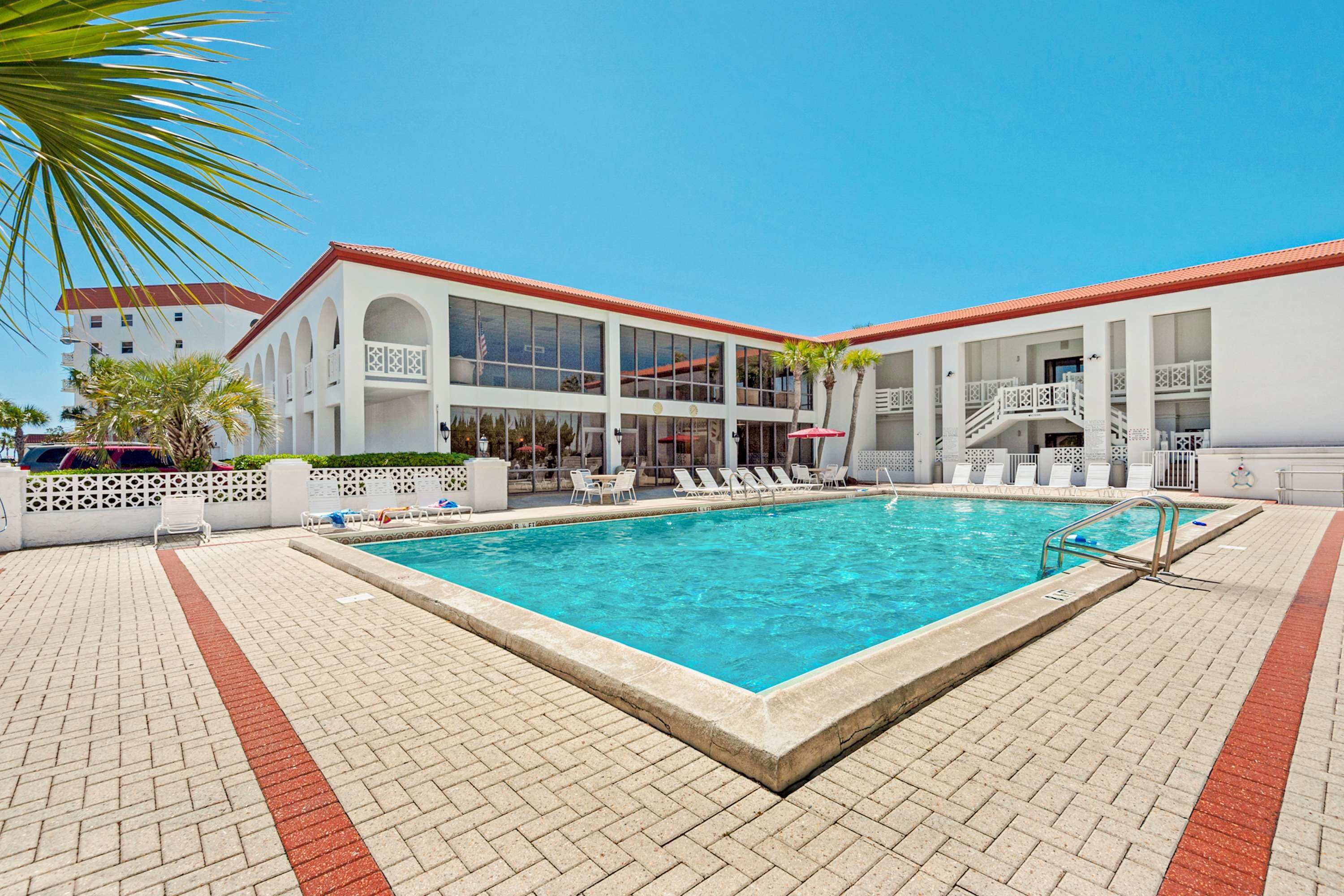 A swimming pool with a tiled deck, white lounge chairs, and a building with balconies and a red roof in the background.