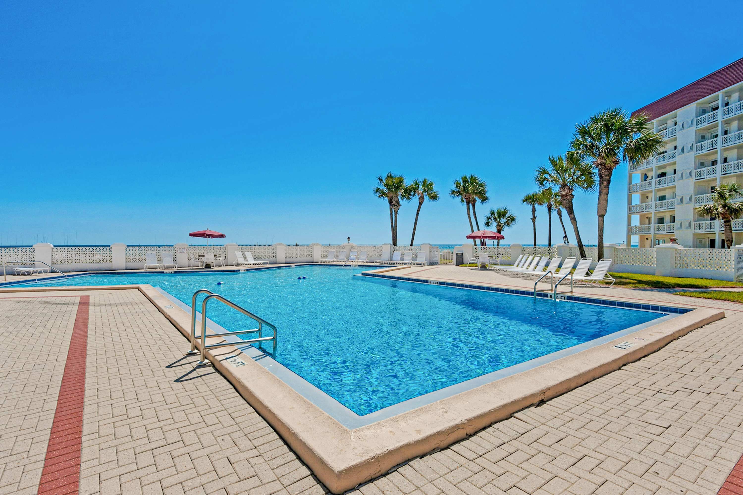 Une piscine avec une terrasse carrelée, des palmiers et un bâtiment doté de balcons en arrière-plan.