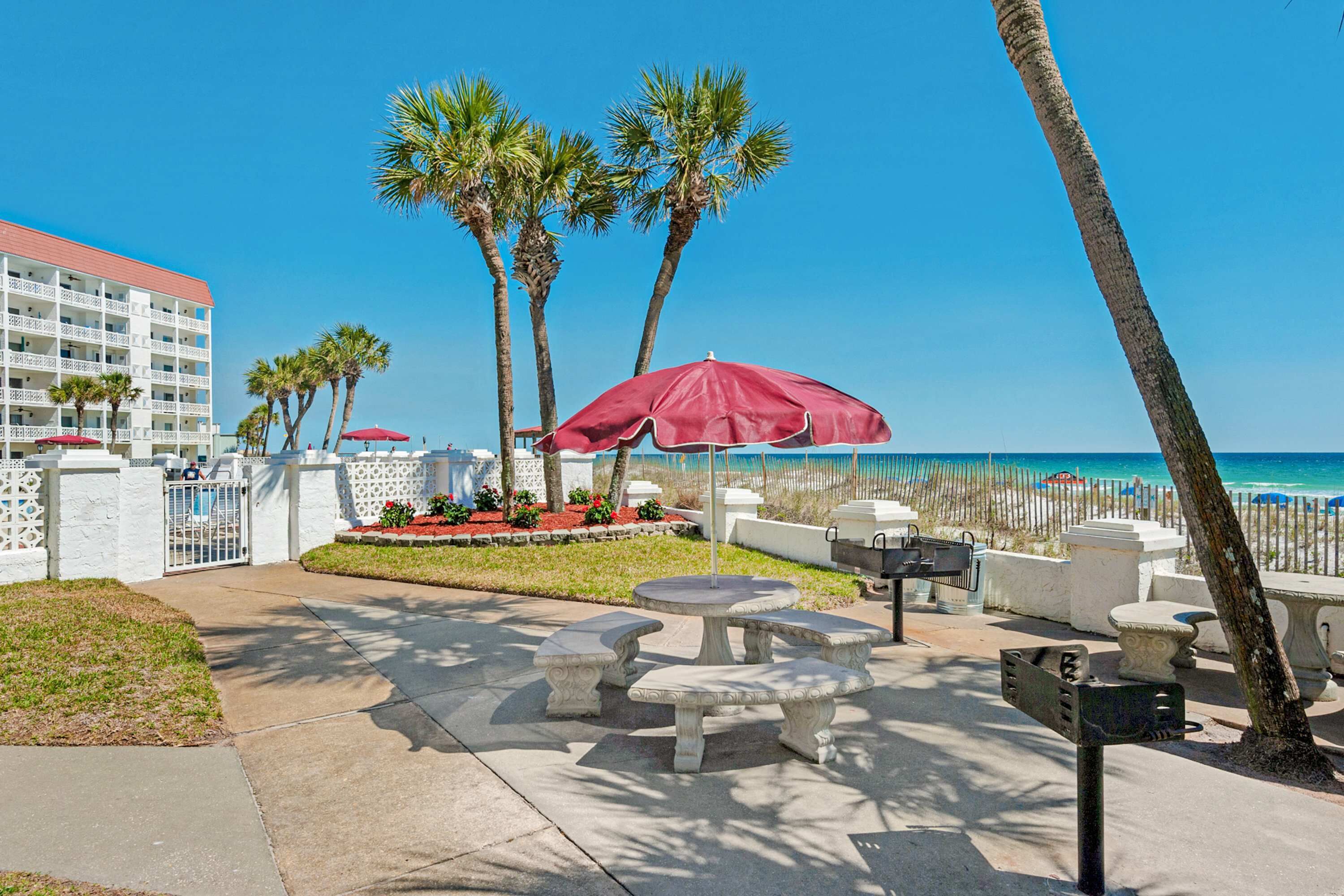 A beachfront patio with stone tables and red umbrellas, palm trees, and a hotel building in the background.