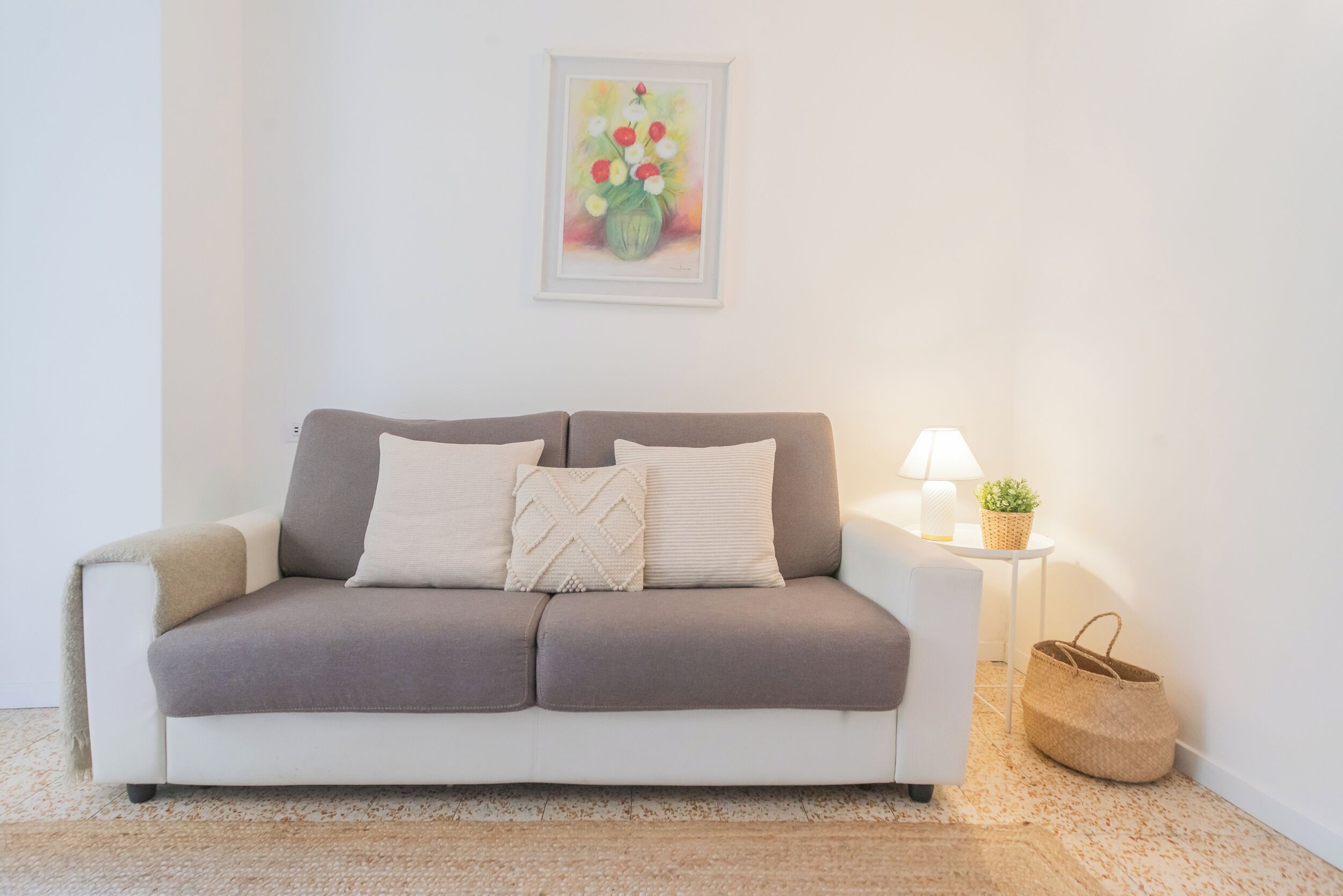 A neutral-toned sofa with a cushion, a small table with a lamp and a plant, and a woven basket on the floor.