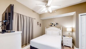 Un dormitorio con cama, ventilador de techo, mesita de noche con lámpara y televisor sobre un mueble.