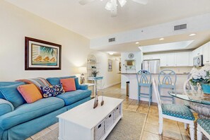 A living room with a blue sofa, a coffee table, and a dining area with a glass table and chairs. - Gorgeous beachfront condo, shared pool and beach access (Fort Walton Beach)