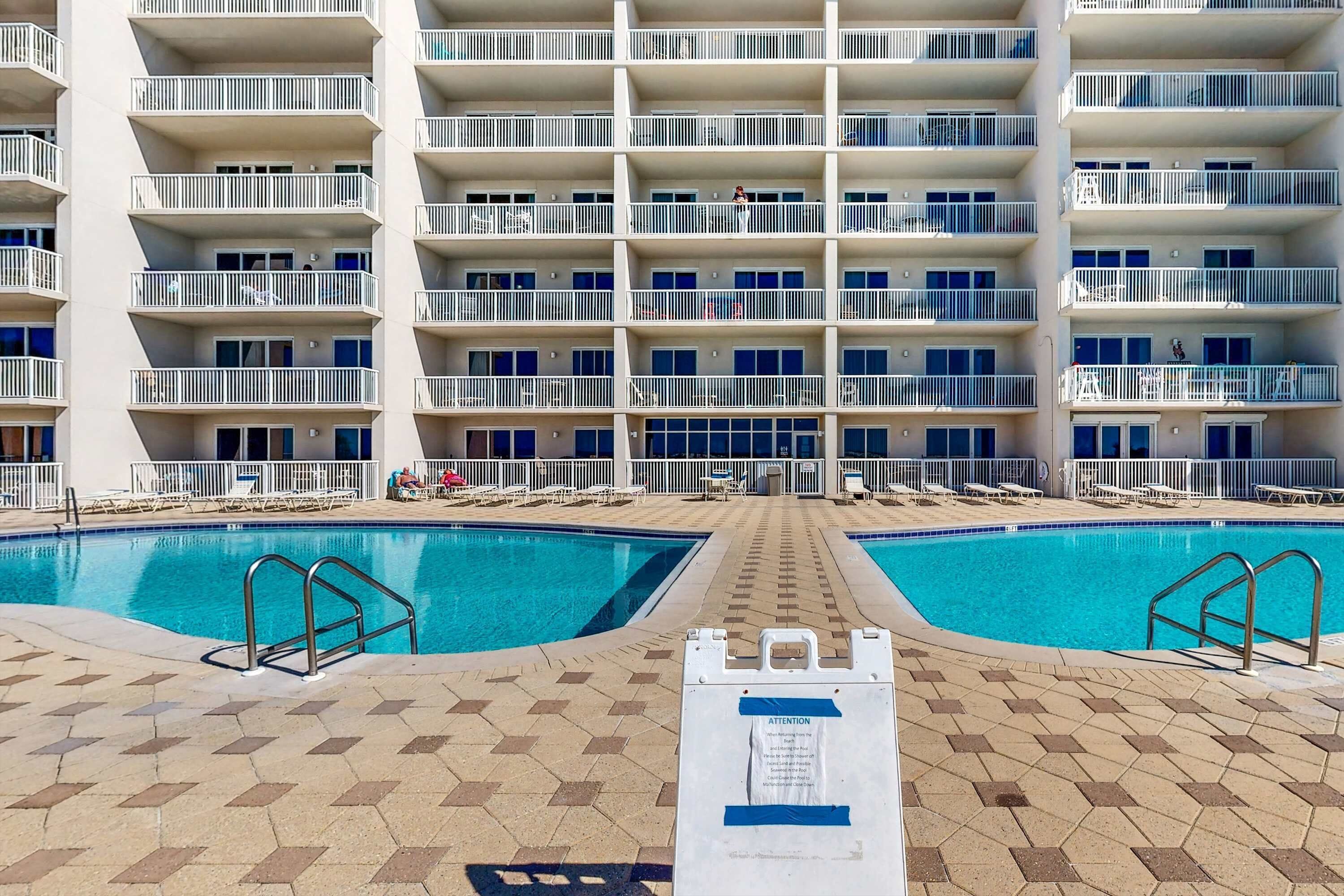 Ein Hotelpoolbereich mit zwei separaten Pools, einem Schild mit einer Hinweis-Aufforderung und einem mehrstöckigen Gebäude mit Balkonen.
