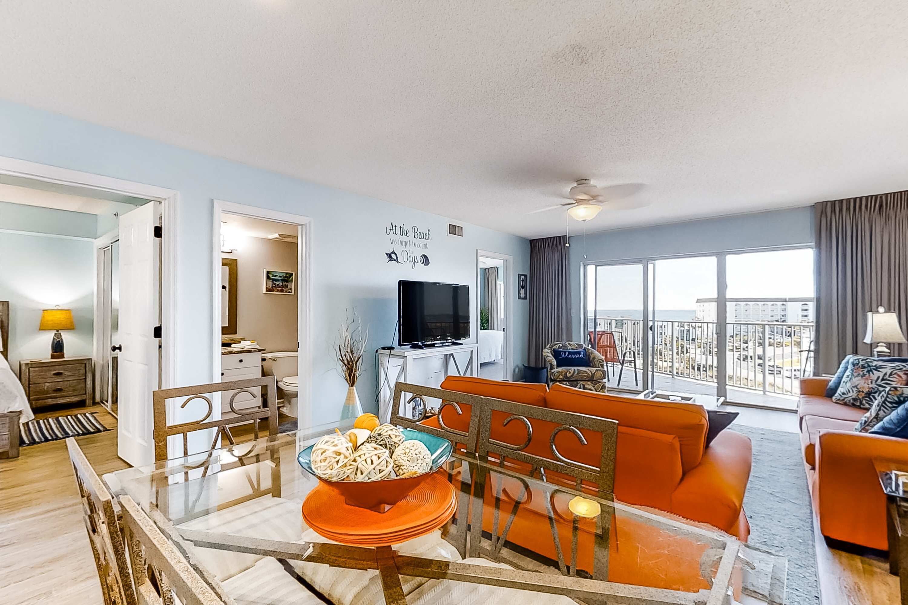 A modern living room with a glass dining table, orange sofas, a TV, and a balcony with a city view.