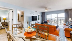 Een moderne woonkamer met een glazen eettafel, oranje banken, een televisie en een balkon met uitzicht op de stad.