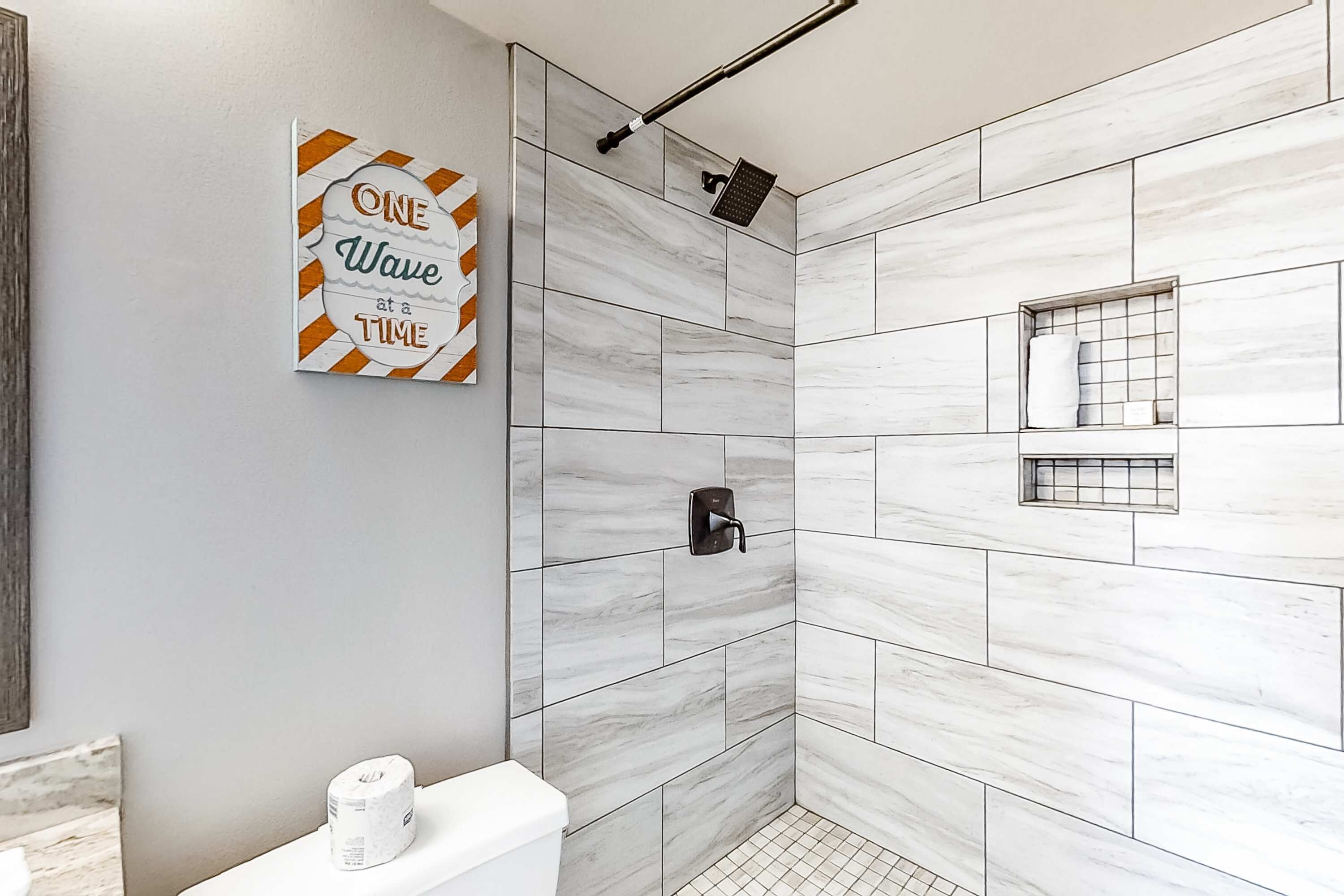 A modern bathroom with white marble tiles, a showerhead, a toilet, and a framed quote on the wall.