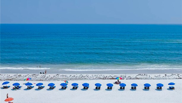 Een strand met rijen blauwe strandstoelen en parasols, een heldere blauwe lucht en de oceaan op de achtergrond.