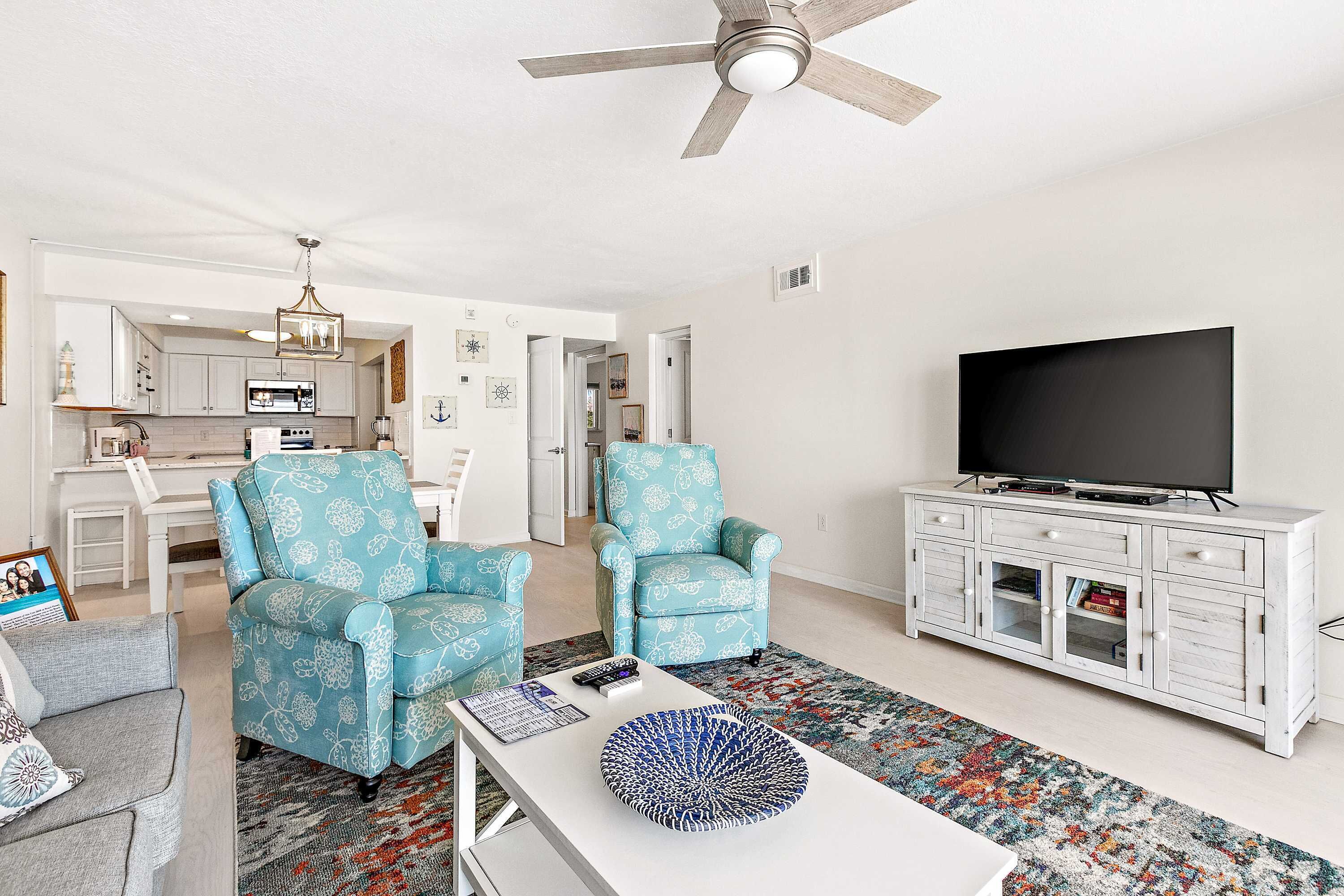 A living room with a TV, two patterned armchairs, a white coffee table, and a kitchen in the background.