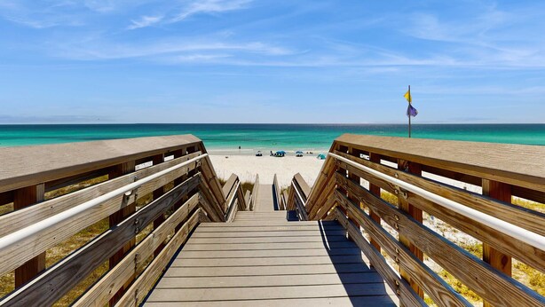 Un paseo marítimo de madera que conduce a una playa con agua turquesa cristalina y una bandera en un asta.