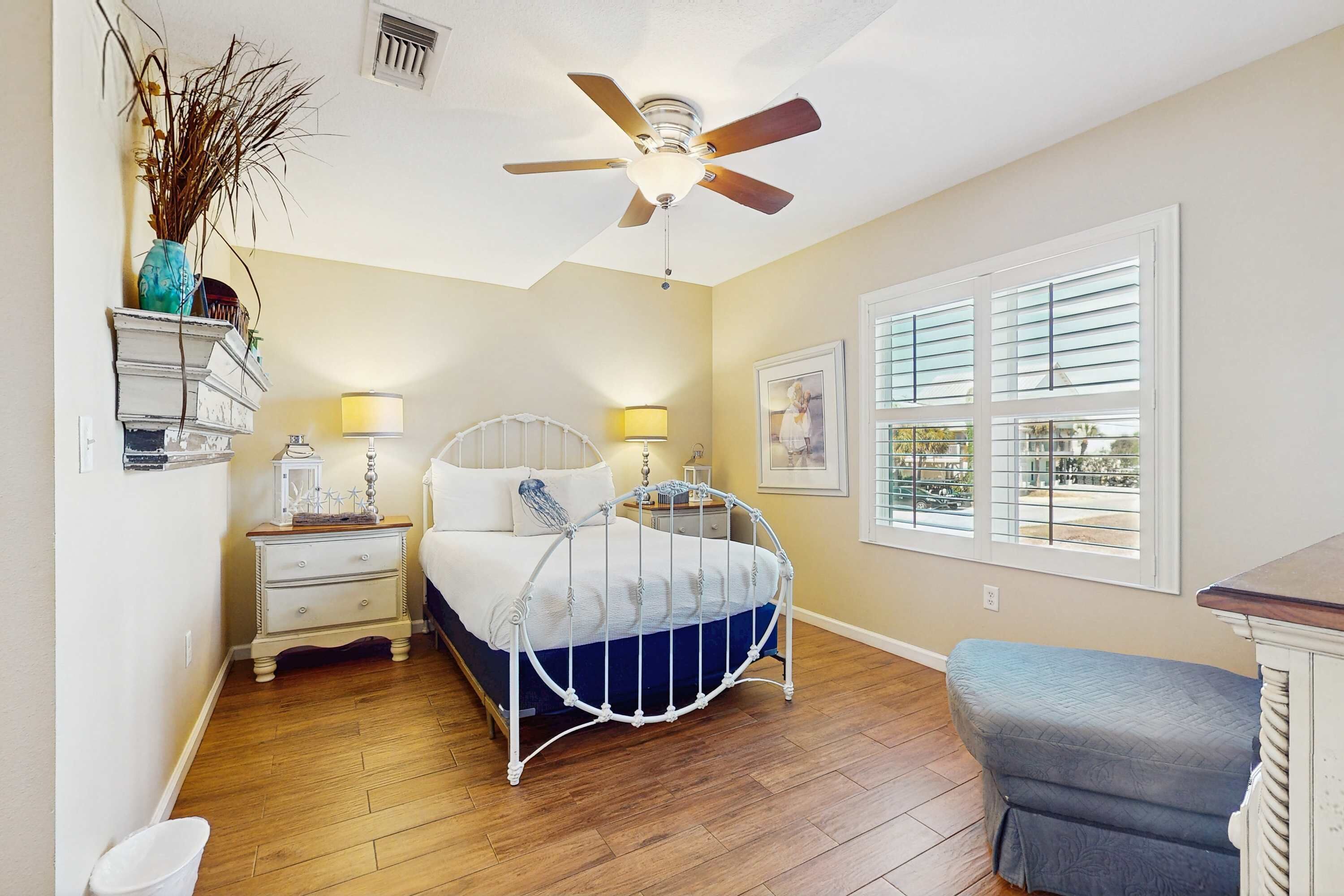 A bedroom with a bed, bedside tables, a nightstand, a ceiling fan, a window with blinds, and a view of the outdoors.