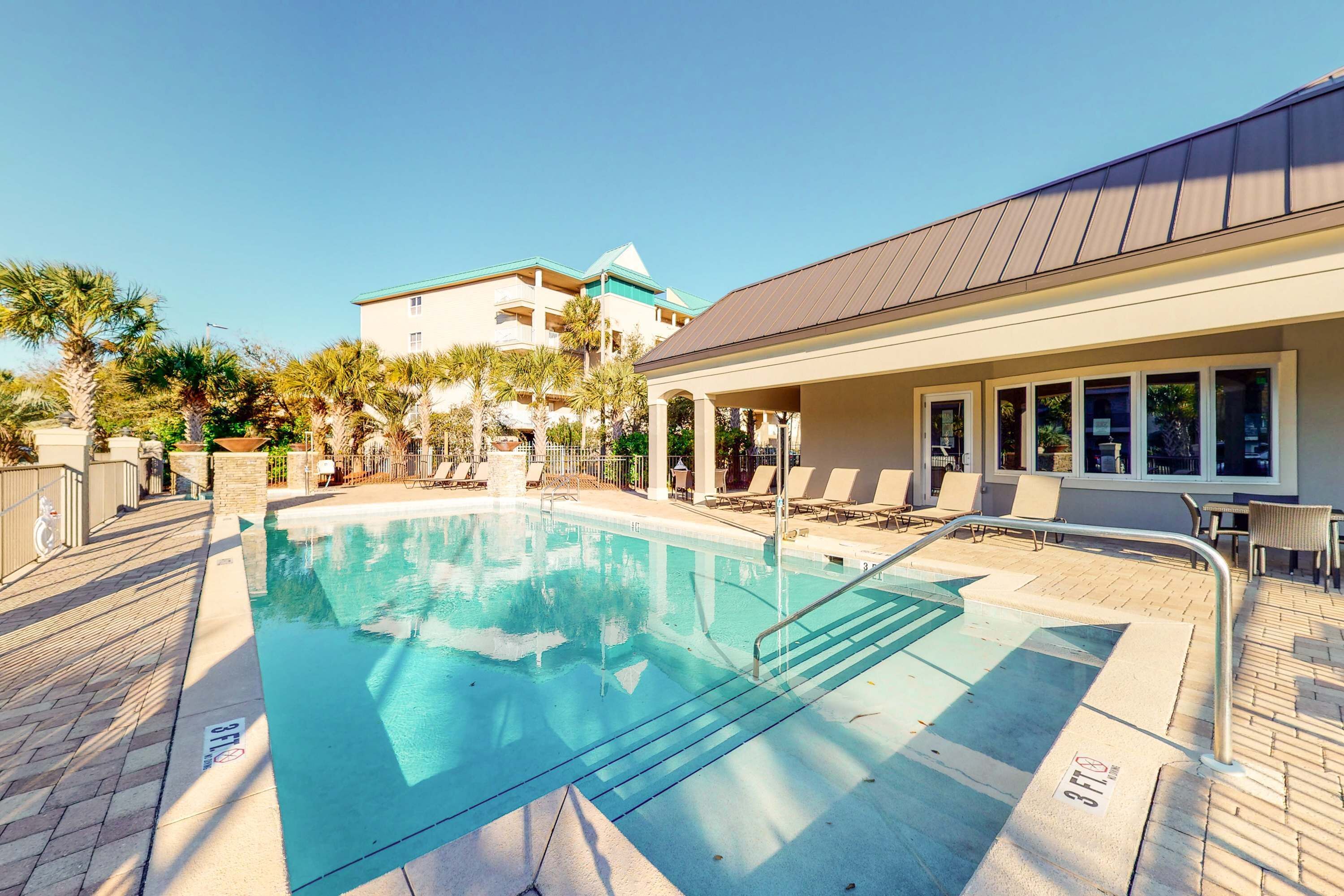A swimming pool with a sunshade area, lounge chairs, and a building with a green roof in the background.