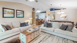Ein geräumiges Wohnzimmer mit zwei Sofas, einem Couchtisch und einem Deckenventilator. Im Hintergrund befindet sich eine Essecke mit einer Chandelierleuchtre und Kunstwerken an den Wänden.
