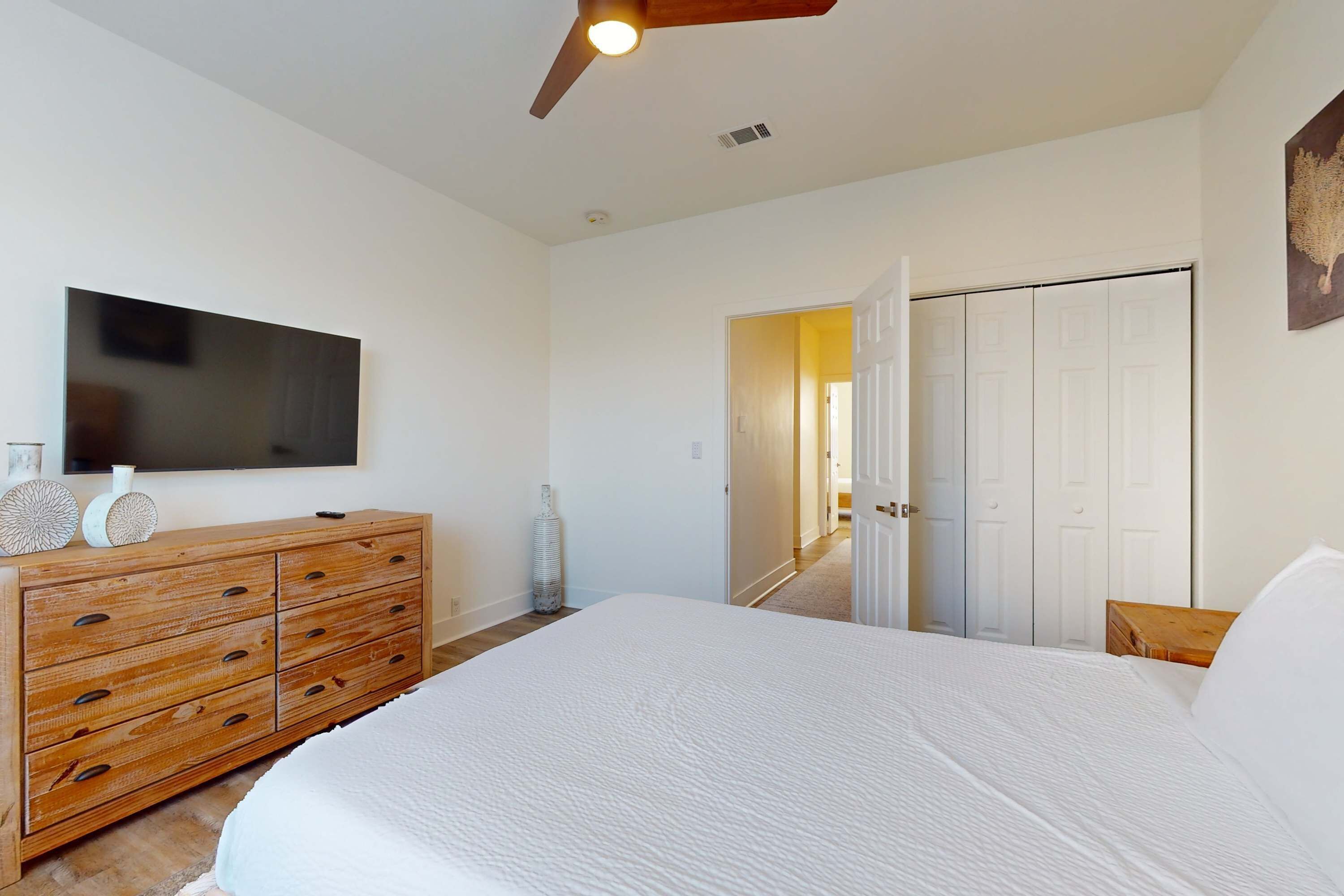 A bedroom with a large bed, wooden dresser, flat-screen TV, and a door leading to another room.