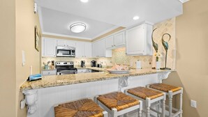 A kitchen with white cabinets, a granite countertop, and a built-in stove. There are three bar stools with woven seats. - Fifth-floor, oceanfront condo with fast WiFi & pool  -  snowbirds OK (Miramar Beach)