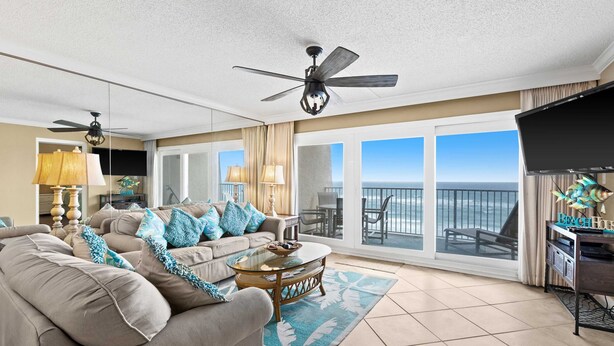 A spacious living room with a large sofa, a coffee table, and a TV. There are ceiling fans, a balcony with a view, and a beach-themed decor. - Fifth-floor, oceanfront condo with fast WiFi & pool  -  snowbirds OK (Miramar Beach)