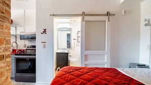 Ein Schlafzimmer mit einer rot gesteppten Bettdecke, einer weißen Tür, einer Küche mit einer Mikrowelle und einer Ziegelwand.