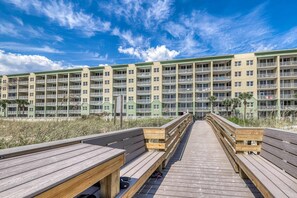 Une promenade en bois mène à un immeuble à plusieurs étages doté de balcons, qui offre une vue sur une plage.