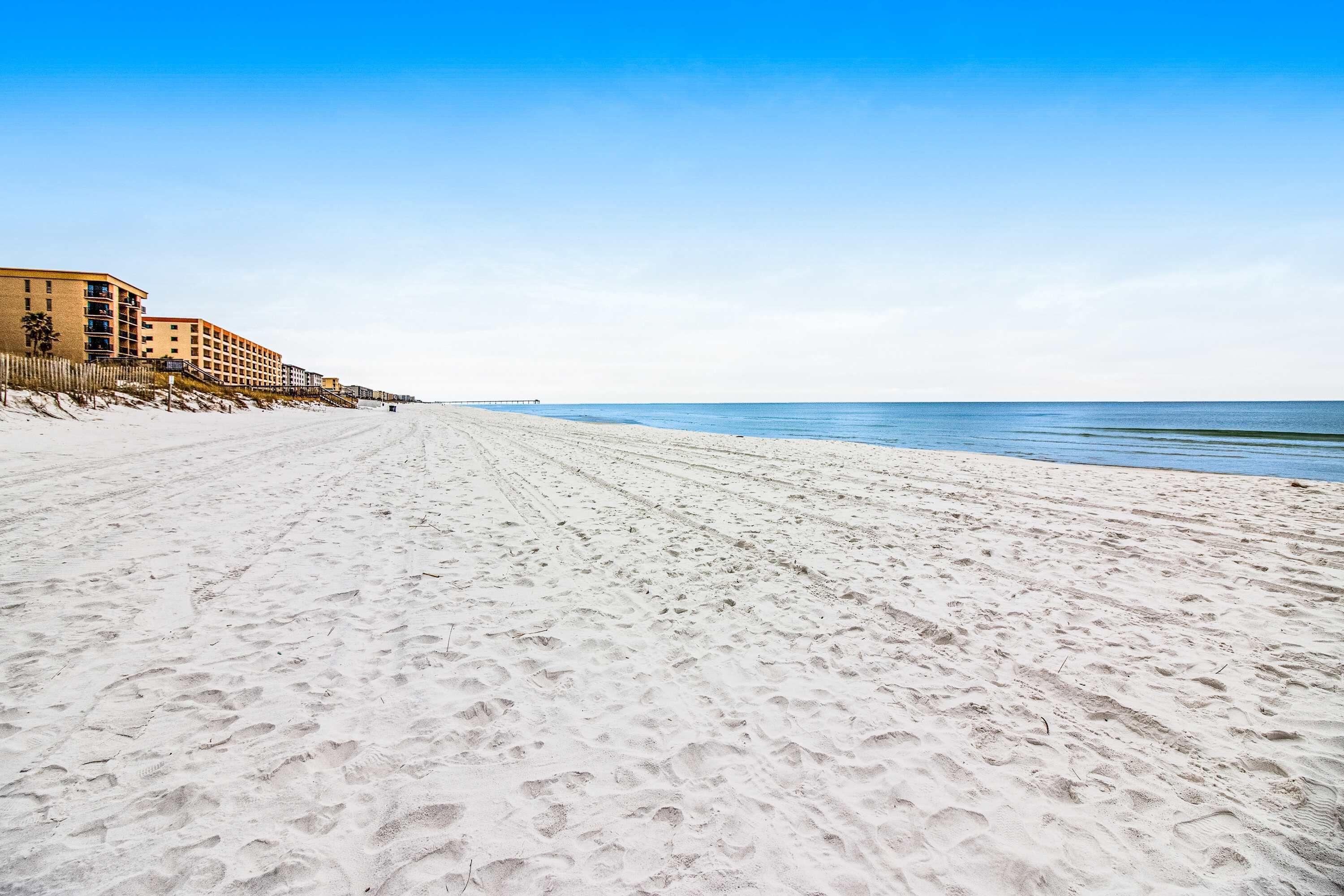 Ein weiter, sandiger Strand mit Fußspuren, die zu einem Mehrstöckiger Gebäude mit Balkonen führen.