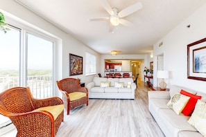 A spacious living room with a white sofa, two wicker chairs, a ceiling fan, and large windows offering a view of the outdoors. - Luxurious Gulf view home w/ a private balcony, shared pool, & on-site golf (Miramar Beach)