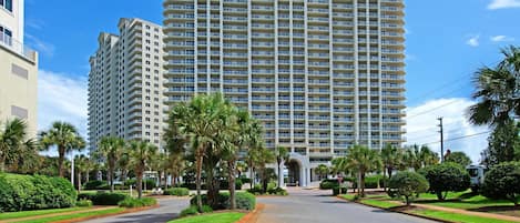 A large, multi-story hotel with a central entrance, surrounded by well-maintained greenery and palm trees.