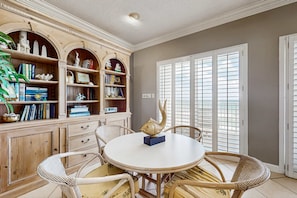 A dining area with a round table, wicker chairs, and a bookshelf with various items. - Oceanfront condo with heated pool, tennis, grilling station, & gym - balcony (Miramar Beach)