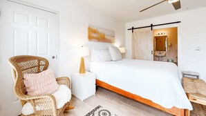 Een slaapkamer met een bed, een stoel, een nachtkastje, een plafondventilator, een wandspiegel en een deur.