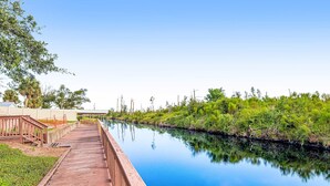 A wooden boardwalk along a calm water body with greenery on one side and a building on the other. - Waterfront Home Close to Beach w/ Shared Pool, Hot Tub, Free WiFi, & Central AC (Mexico Beach)