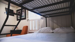 Un lit superposé avec une literie blanche, un bureau avec une chaise et une fenêtre avec des volets.