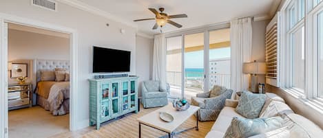 A spacious living room with a ceiling fan, a flat-screen TV, a coffee table, and a sofa. There is a balcony with a view of the ocean.