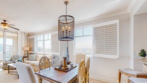 Amplio salón con mesa de comedor de madera, muebles blancos y ventanas grandes con persianas.