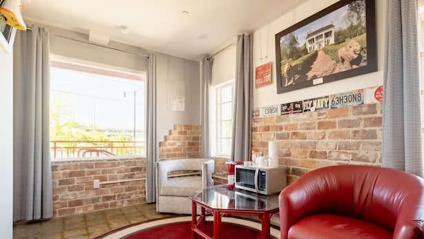 A room with a brick wall, a red leather chair, a white armchair, a small table with a microwave, and a framed picture on the wall. - Alabama Boutique Hotel: #6 w/ TV Rm, PacMan Arcade (Tuscaloosa)