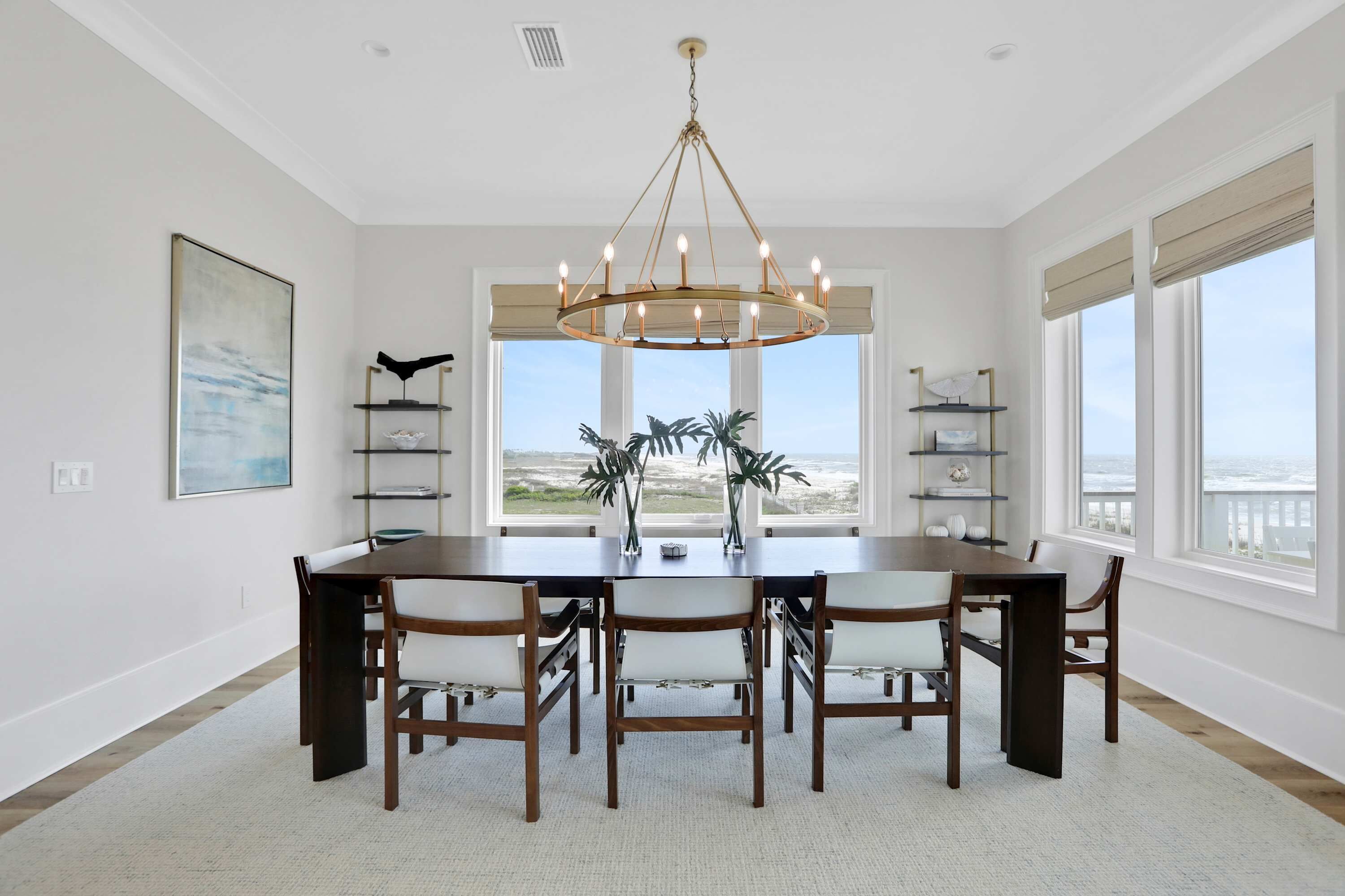 Une salle à manger avec une grande table, six chaises, un lustre et une vue sur l’océan grâce à de grandes fenêtres.
