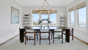 A dining room with a large table, six chairs, a chandelier, and a view of the ocean through large windows. - Newly constructed beachfront home with panoramic bay & Gulf views (St. George Island)