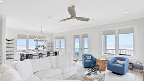 A spacious living room with a white sofa, a blue armchair, and a dining area with a view of the ocean. - Newly constructed beachfront home with panoramic bay & Gulf views (St. George Island)