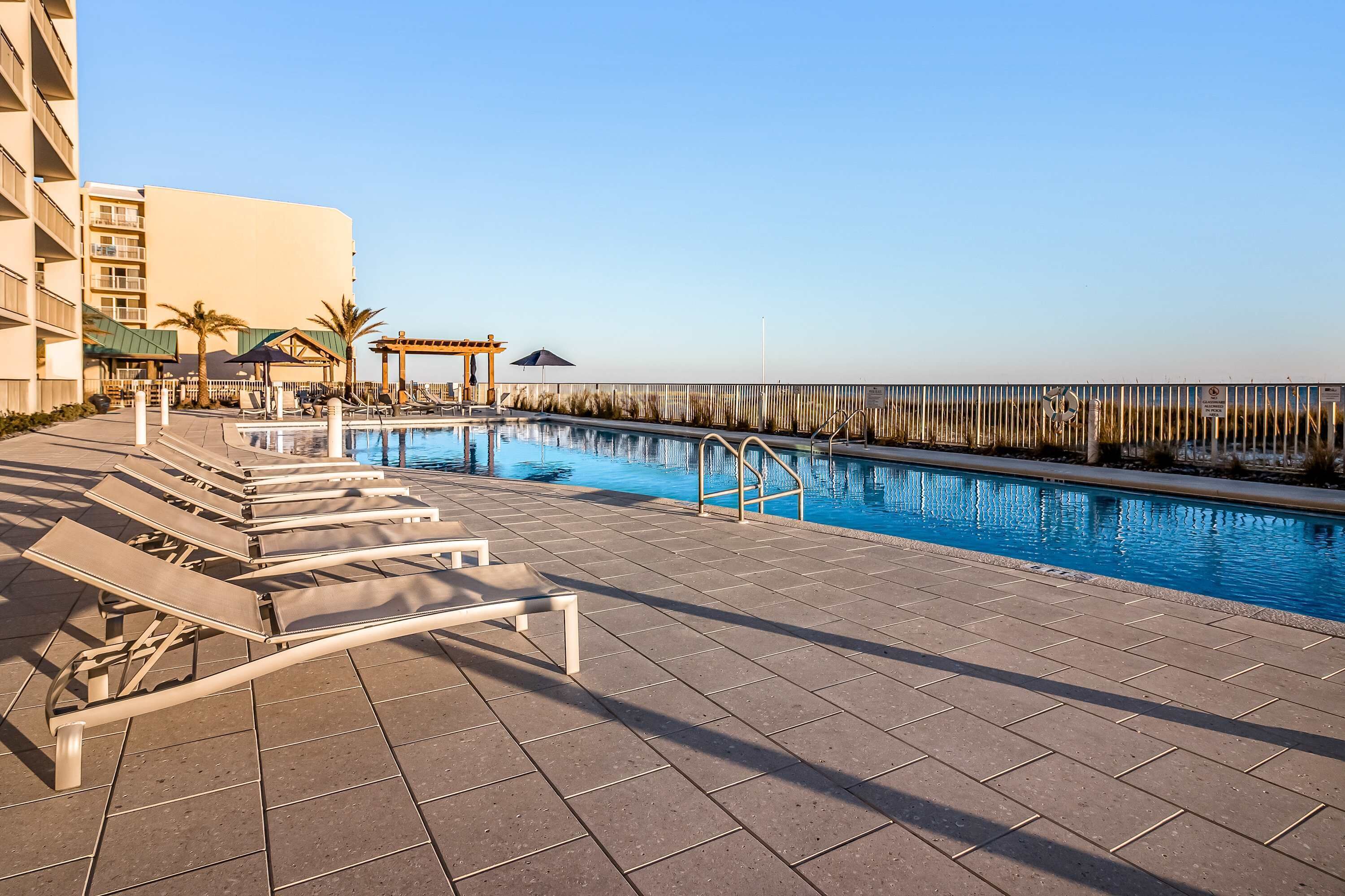 A swimming pool with lounge chairs and a pergola, surrounded by a fence and a building.