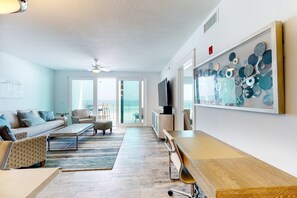Ein modernes Wohnzimmer mit Sofa, Couchtisch und Fernseher. Ein großes Fenster bietet Blick ins Freie.