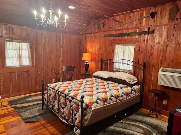 A wooden-paneled bedroom with a bed, a chandelier, a lamp, and a heater.