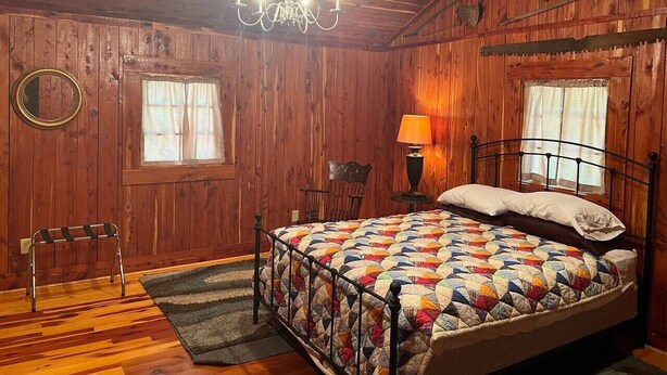 Un dormitorio con paneles de madera, una cama, una araña, una silla y una pequeña mesa con lámpara.