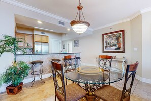 A dining area with a glass table and chairs, a hanging light fixture, and a potted plant. - Gulf Front 4BR w Balcony, Pool, Hot Tub & W&D (Fort Walton Beach)