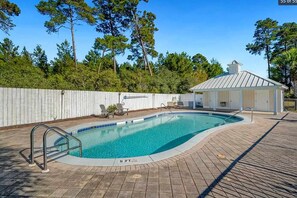 Ein Schwimmbecken mit Holzterrasse, überdachter Sitzgelegenheit und weißem Zaun.