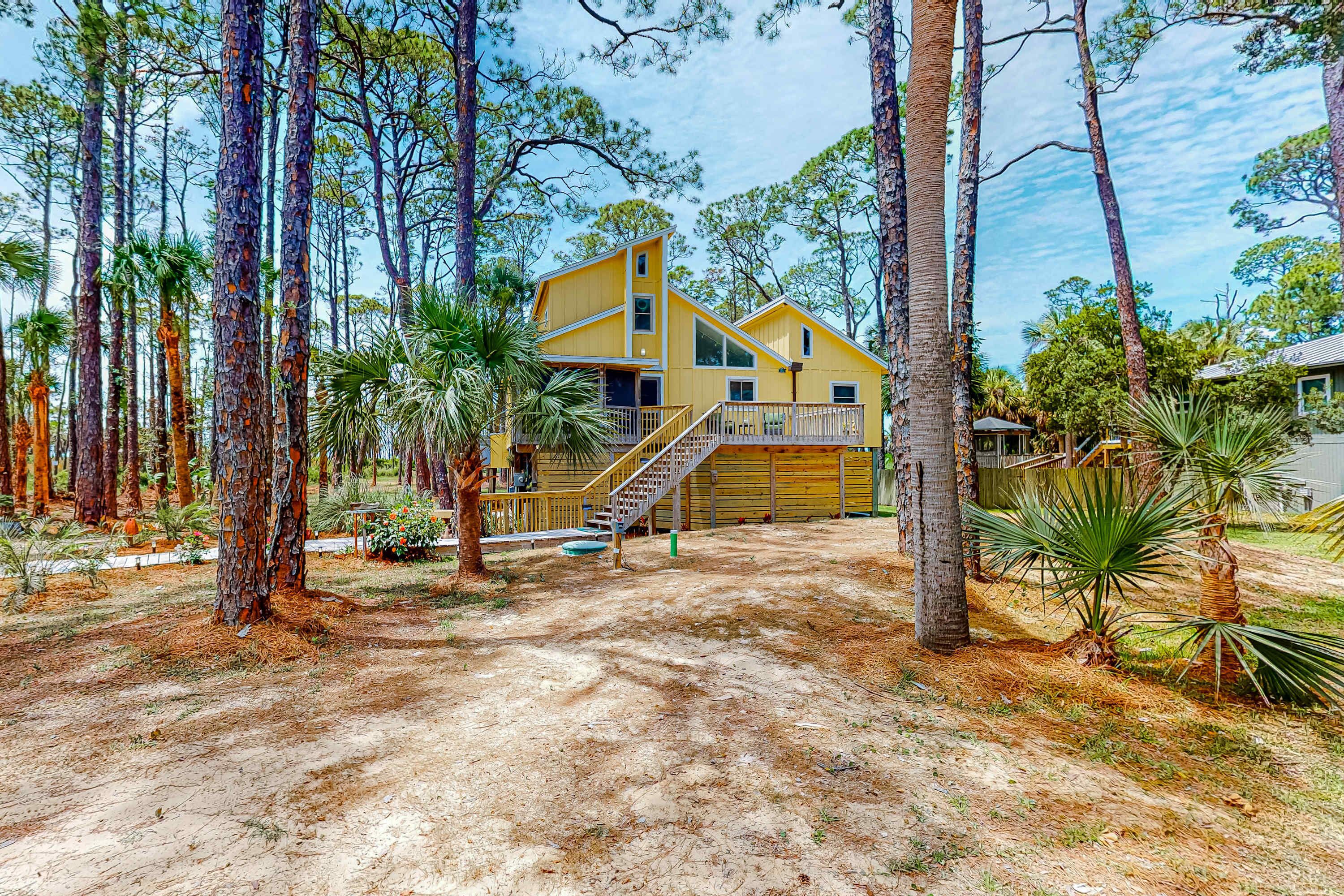 A two-story yellow house with a staircase leading to a balcony, surrounded by tall pine trees and palm plants.
