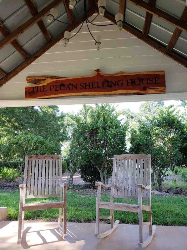 A wooden sign reading "The Pecan Sheltering House" above two rocking chairs in a covered outdoor area with a view of trees.