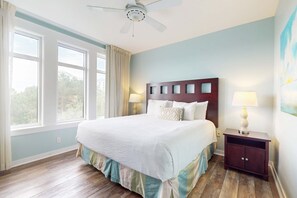 Quarto com cama grande, mesa de cabeceira, ventilador de teto e janela com cortinas.