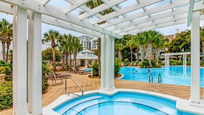 Área da piscina com pérgola, palmeiras e um edifício ao fundo.