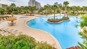 A resort pool area with multiple lounge chairs, a central pool, and surrounding palm trees.
