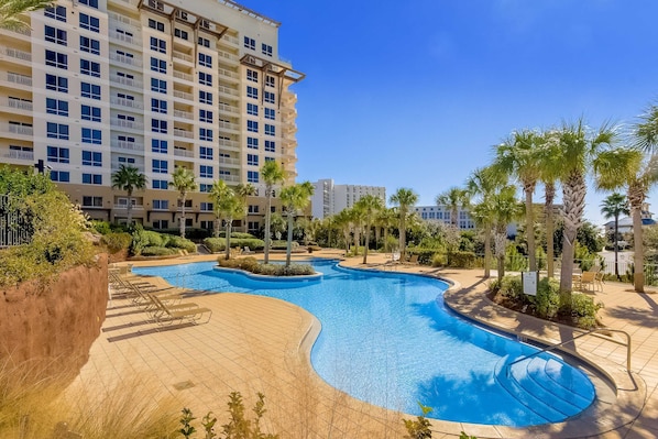 A swimming pool with a curved design, surrounded by palm trees and lounge chairs, with a multi-story building in the background.
