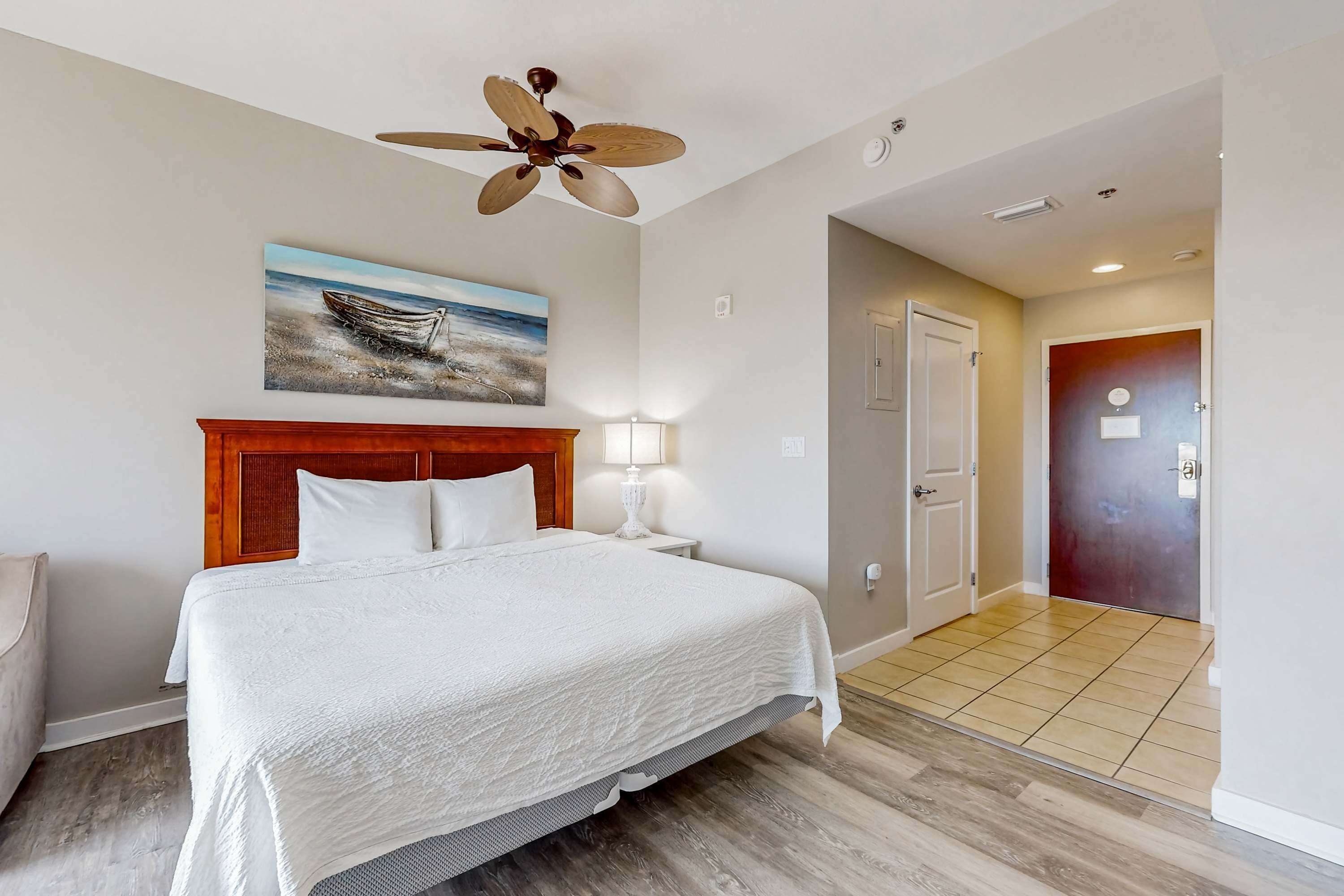 Una habitación de hotel con cama, ventilador de techo, un cuadro de un barco en la pared, una mesita de noche con lámpara y una puerta al pasillo.