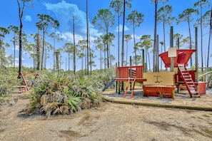 A playground with a slide and climbing structures, surrounded by tall pine trees.