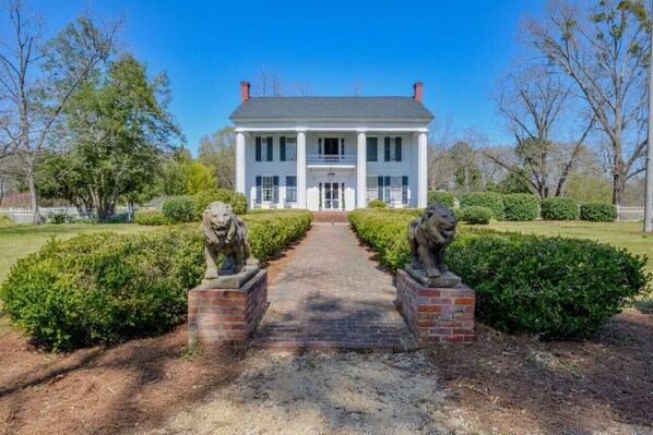 A large, stately house with a central entrance flanked by two lion statues, surrounded by well-maintained hedges and trees.