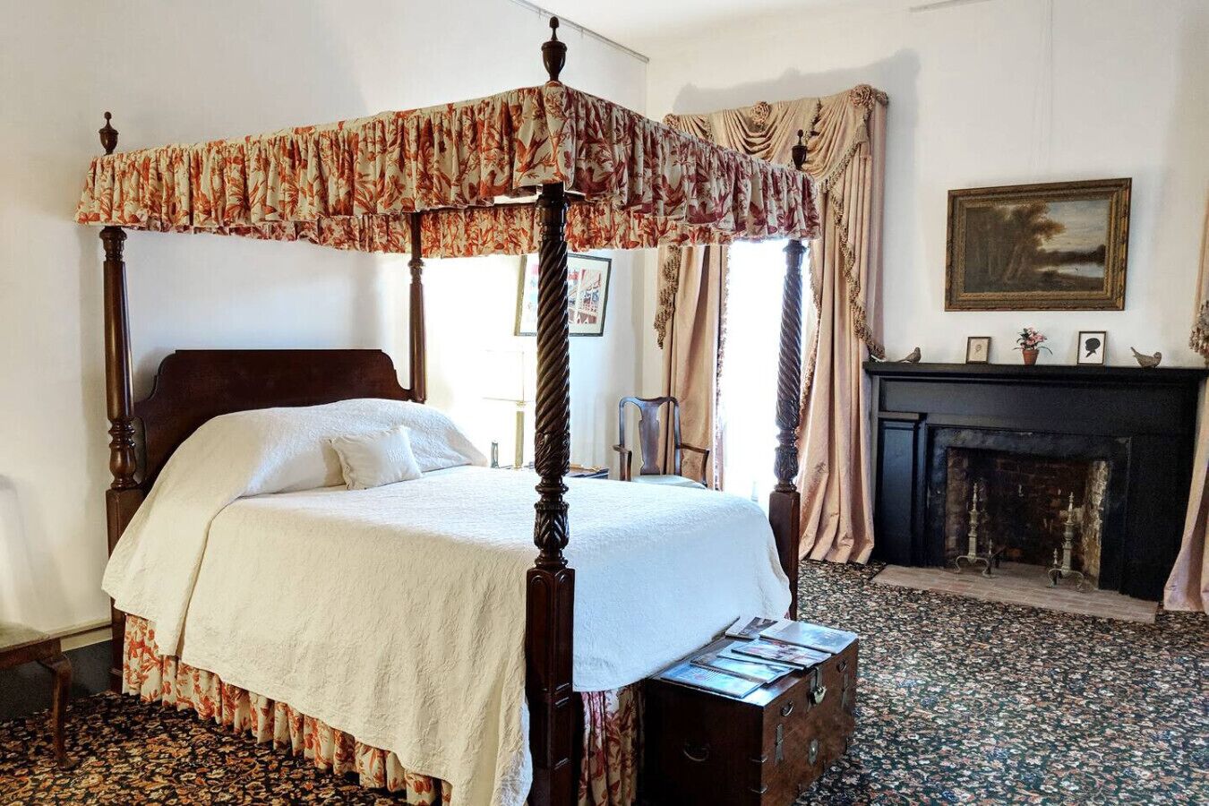 Ein Bett mit Baldachin und Blumenmuster, ein Kamin mit gerahmten Bildern und ein gemustertes Teppich.
